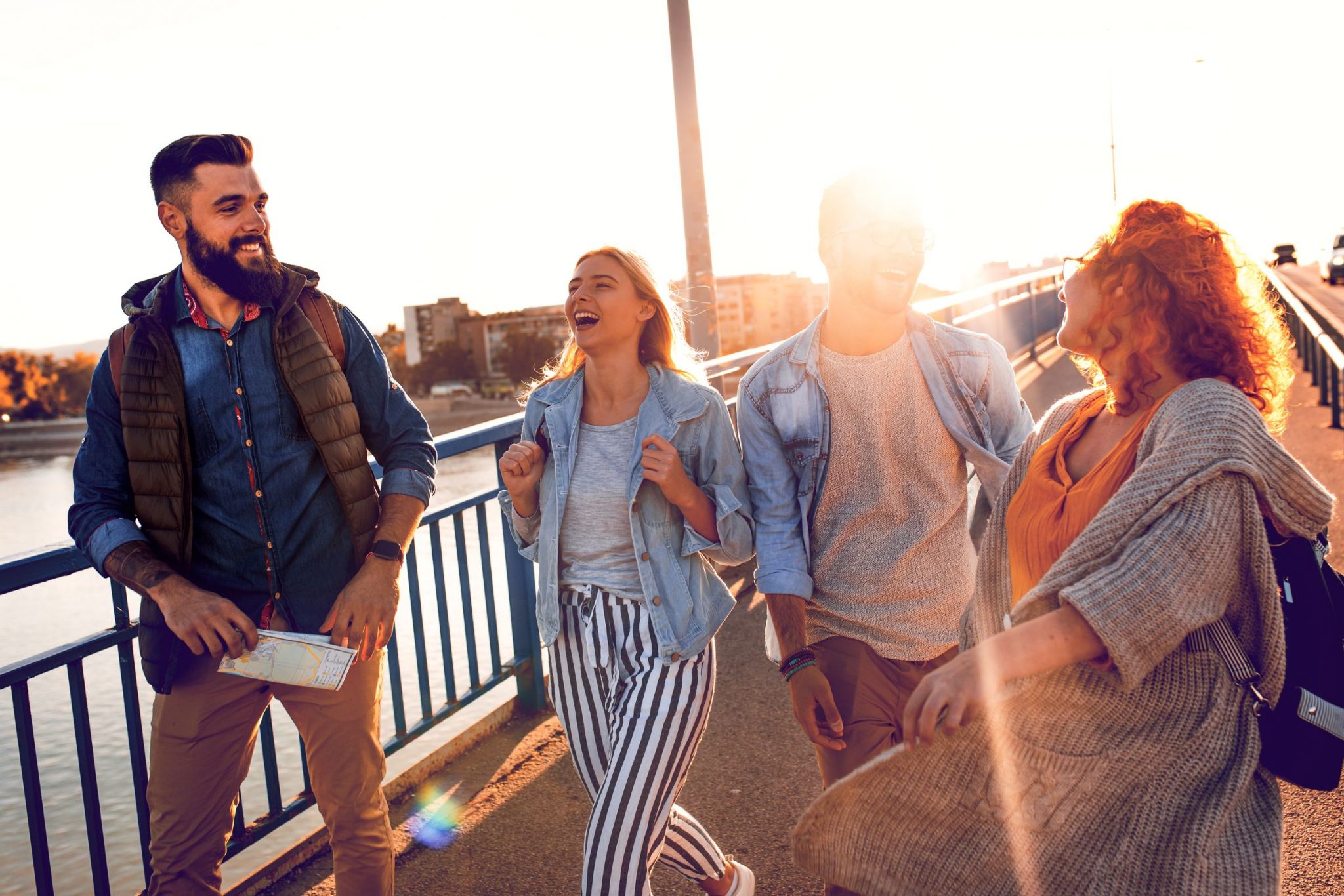 A group of four friends is walking and laughing on a bridge in Chattanooga during a sunny day, with buildings and water in the background.