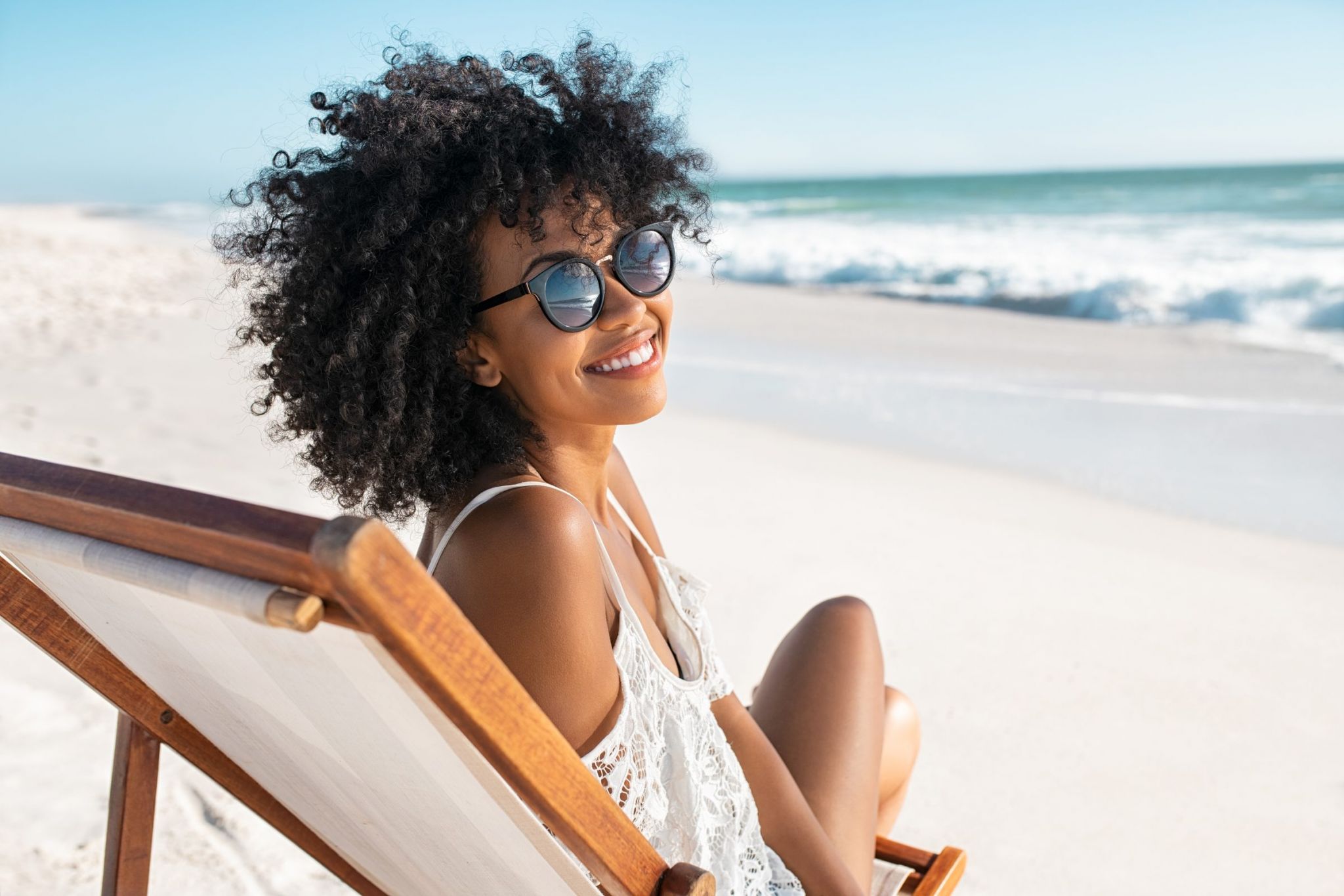 A happy young woman relaxing on a wooden deck chair resting on a sandy beach along the ocean shore